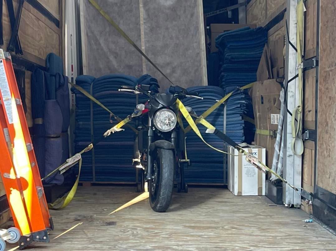 Motorcycle strapped with yellow tie-downs inside a truck or storage trailer, facing forward.