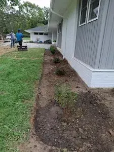 A newly dug garden bed next to a house with visible plants, two people working in the background.