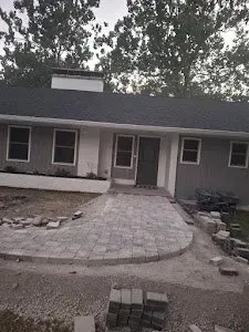 Home exterior with paving stones under construction. Gray walls, black roof, trees in background.