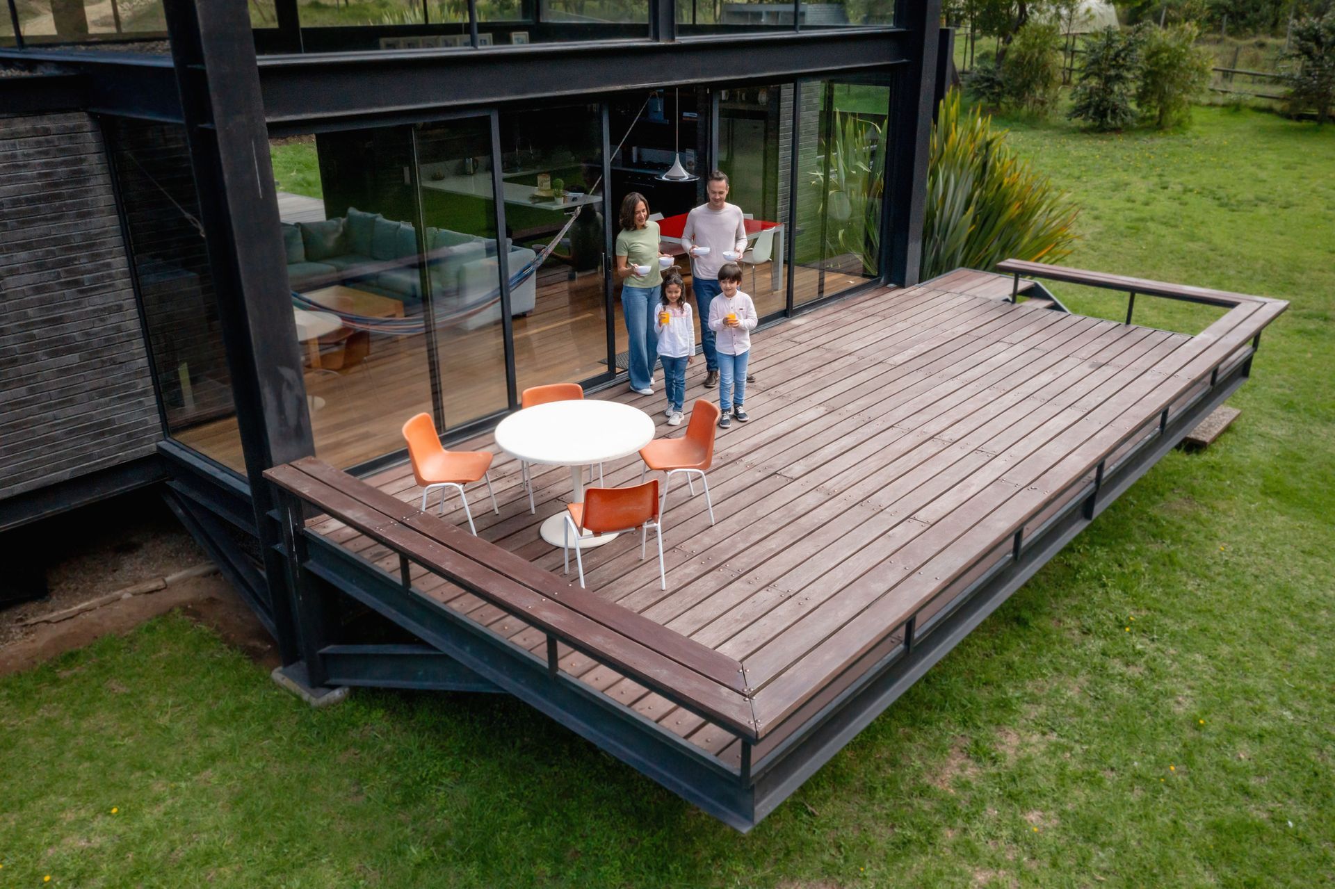 Family of four on a wooden deck in front of a modern house, with chairs and table, green grass surrounding.