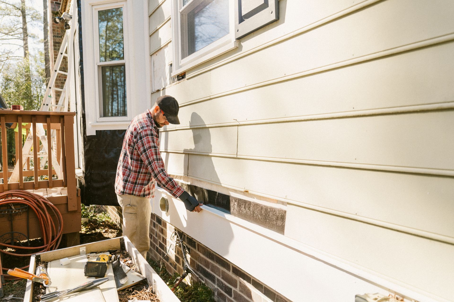 Man installing siding on a house exterior; tan, brick, and cream colors.
