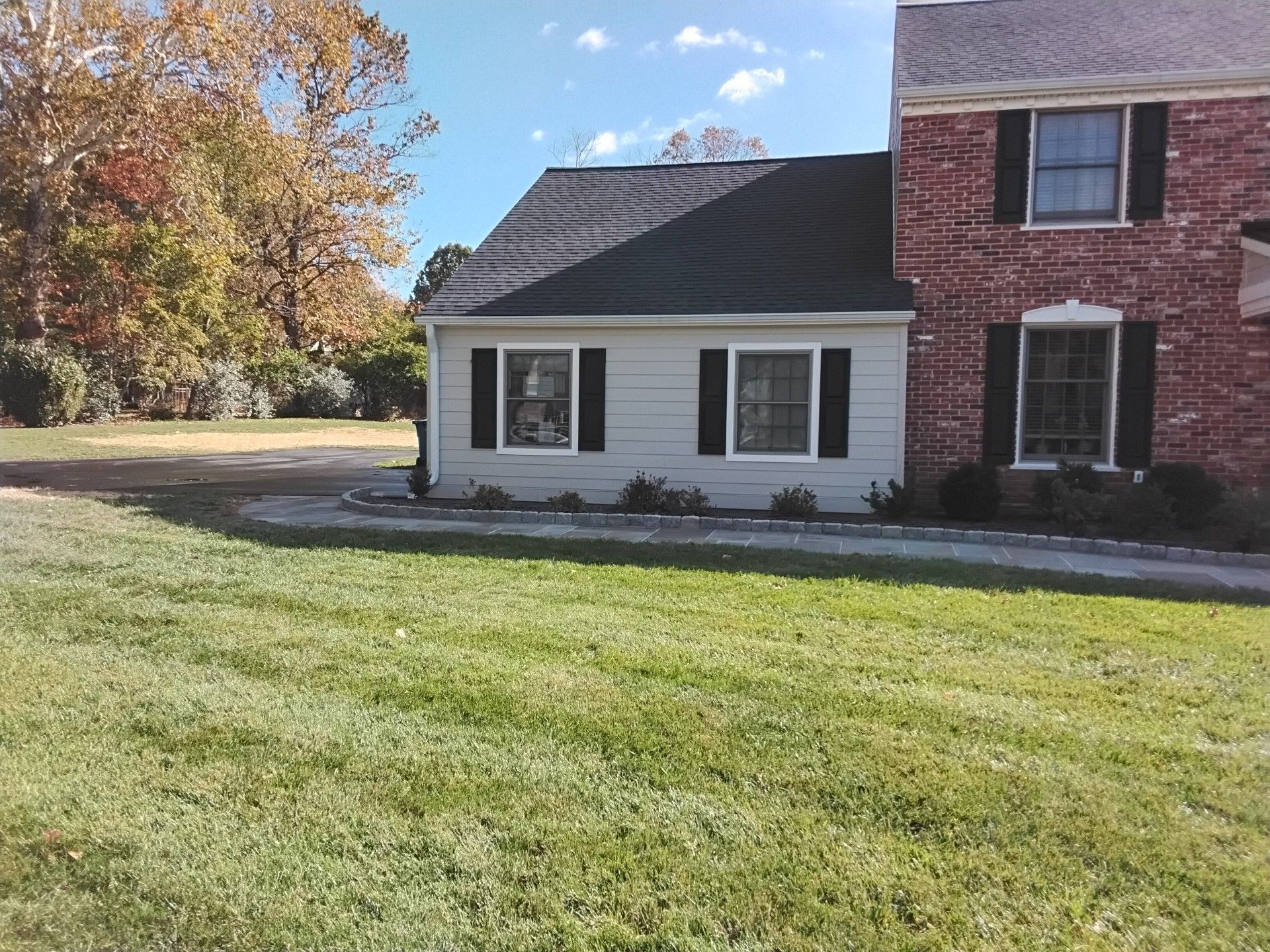 House with a white-sided addition, black shutters, and red brick siding on the right. Green lawn and trees in the background.