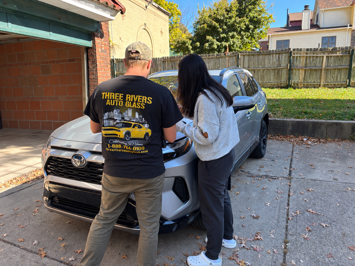 Two people stand beside a silver car in a driveway, inspecting the front end.