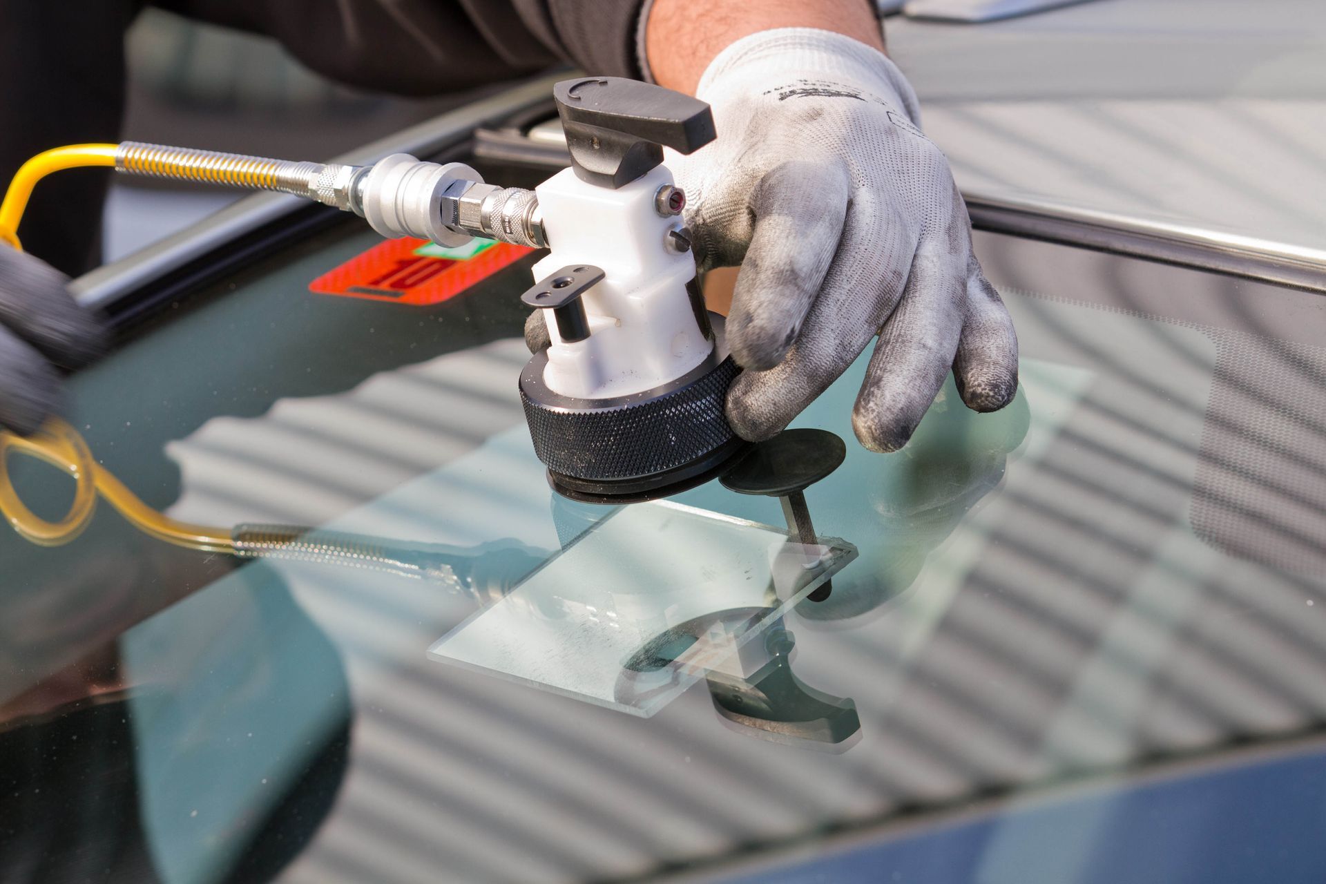 Gloved hand using a polishing tool on a car windshield, with yellow cords and reflections visible