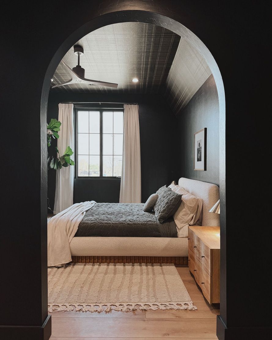 A modern bedroom viewed through a black arched doorway, featuring black walls, a light rug, and a textured ceiling.