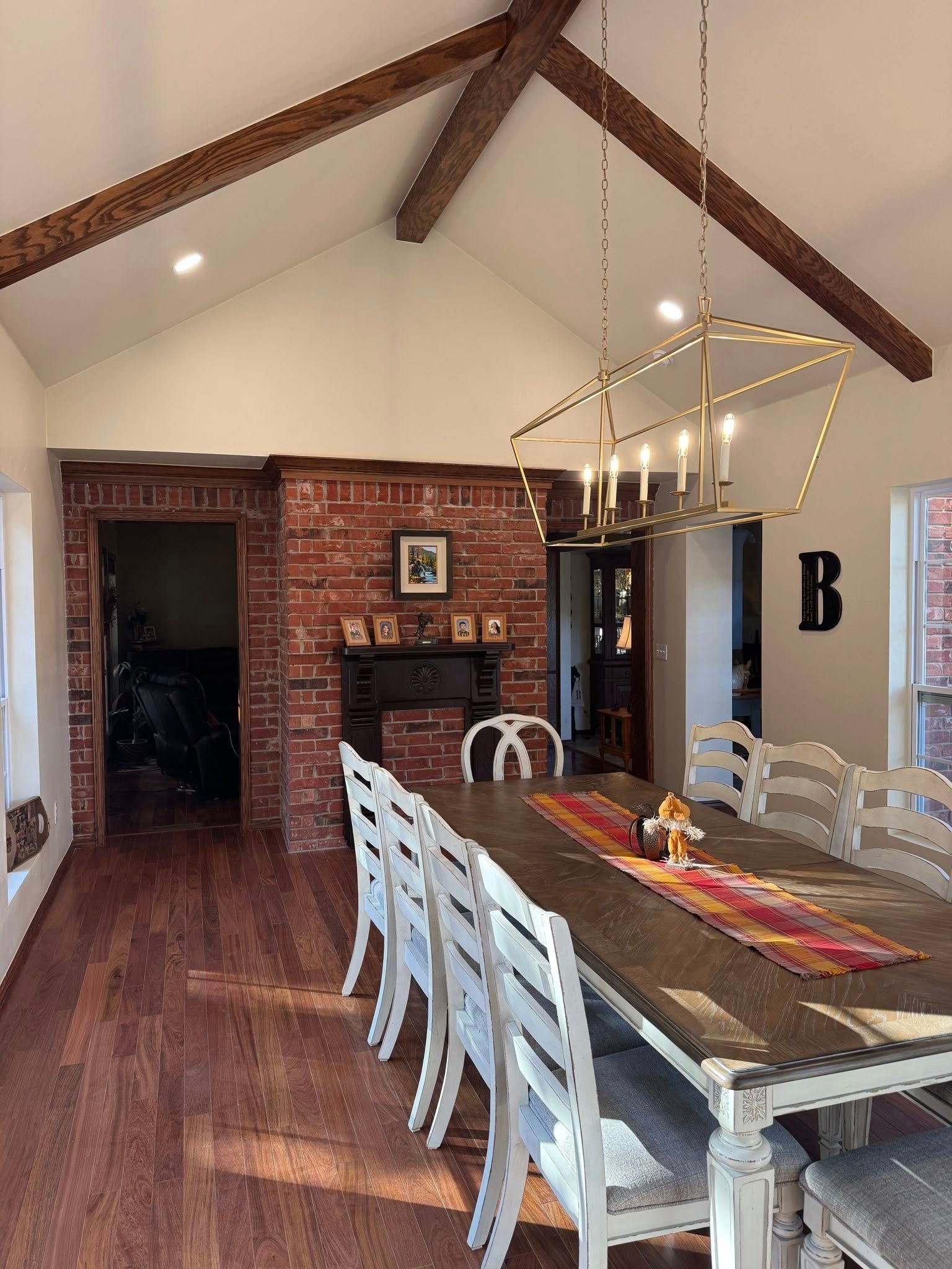 Dining room with vaulted ceiling, exposed wooden beams, brick wall, long table, white chairs, and a geometric chandelier.