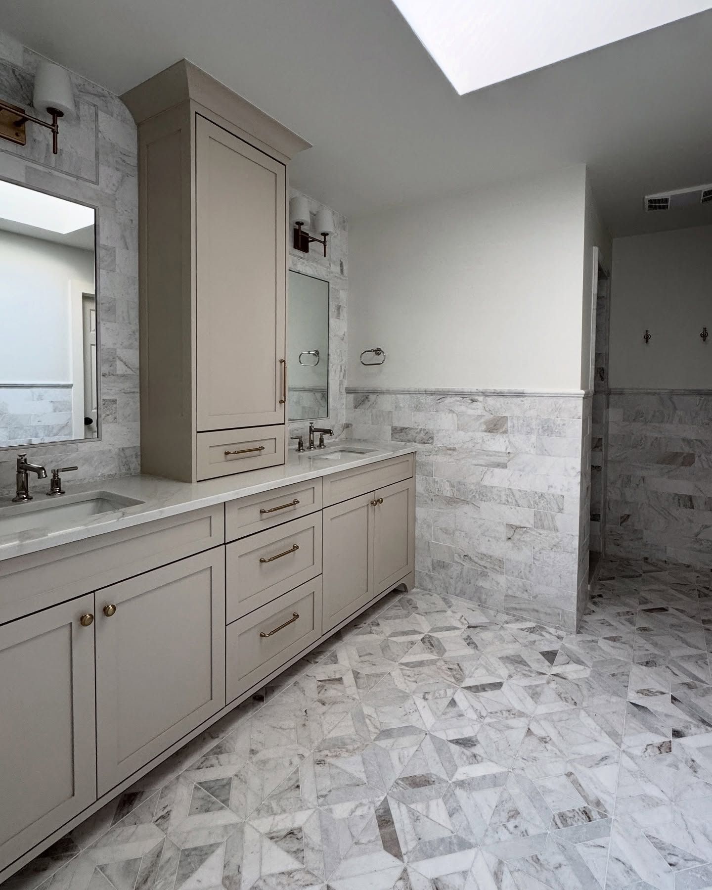 A bathroom with a taupe double vanity, marble tile floor and walls, a skylight, and wall-mounted vanity lights.
