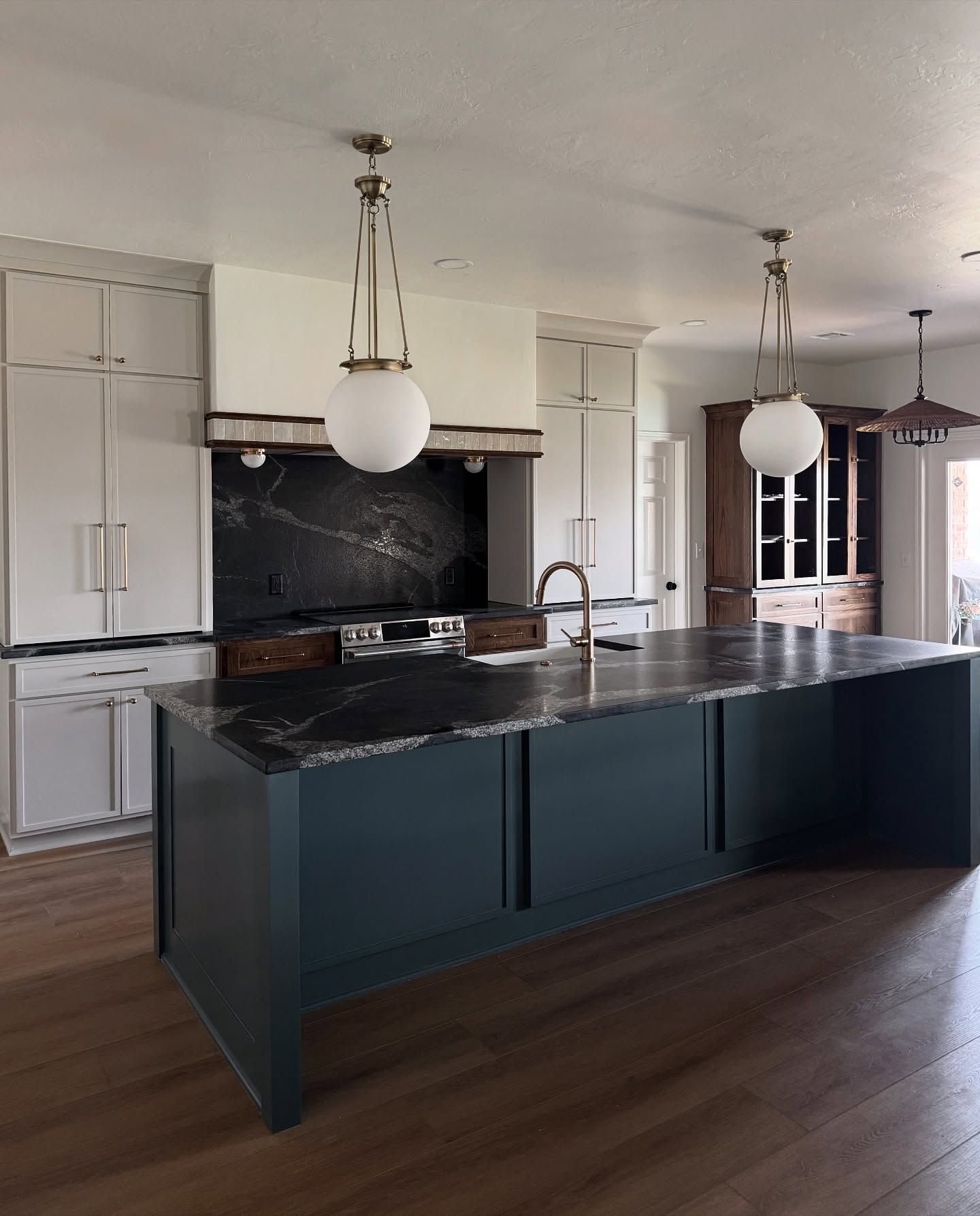 A modern kitchen with a large, dark navy island, white cabinetry, a black marble backsplash, and hanging globe lights.