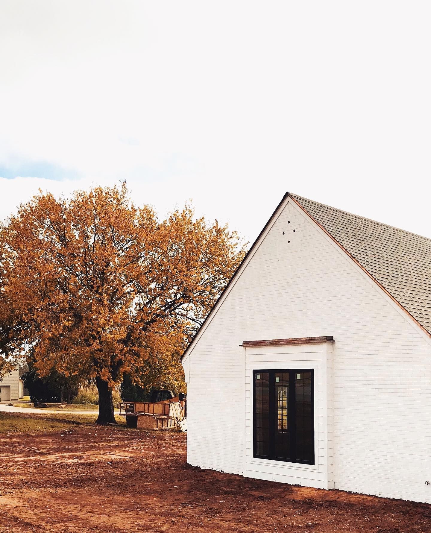 A white-painted brick building with a black-framed window, next to a large autumn tree with orange leaves.