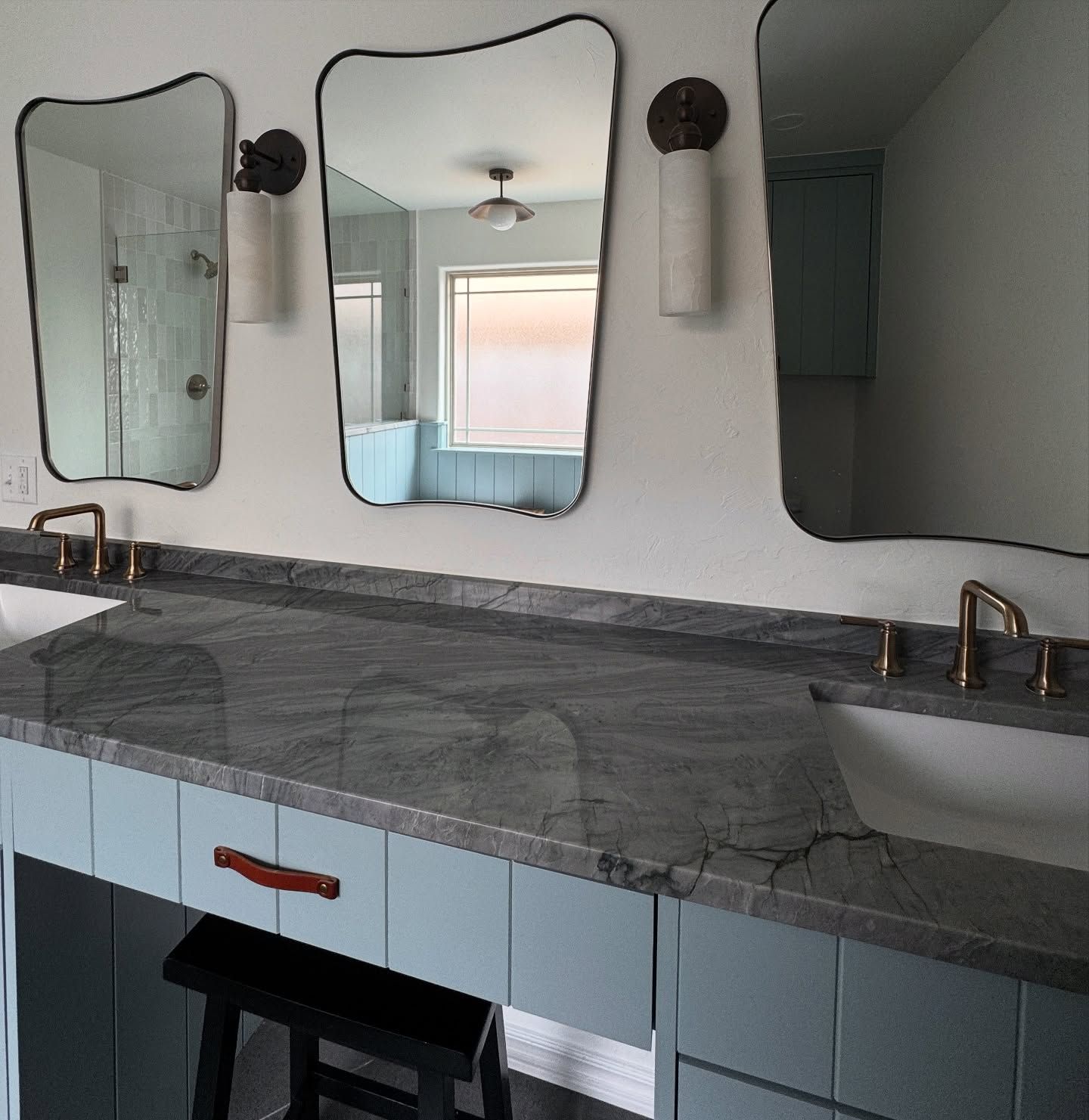 A bathroom vanity with three asymmetrical mirrors, light blue cabinetry, leather drawer pulls, and dark stone countertops.