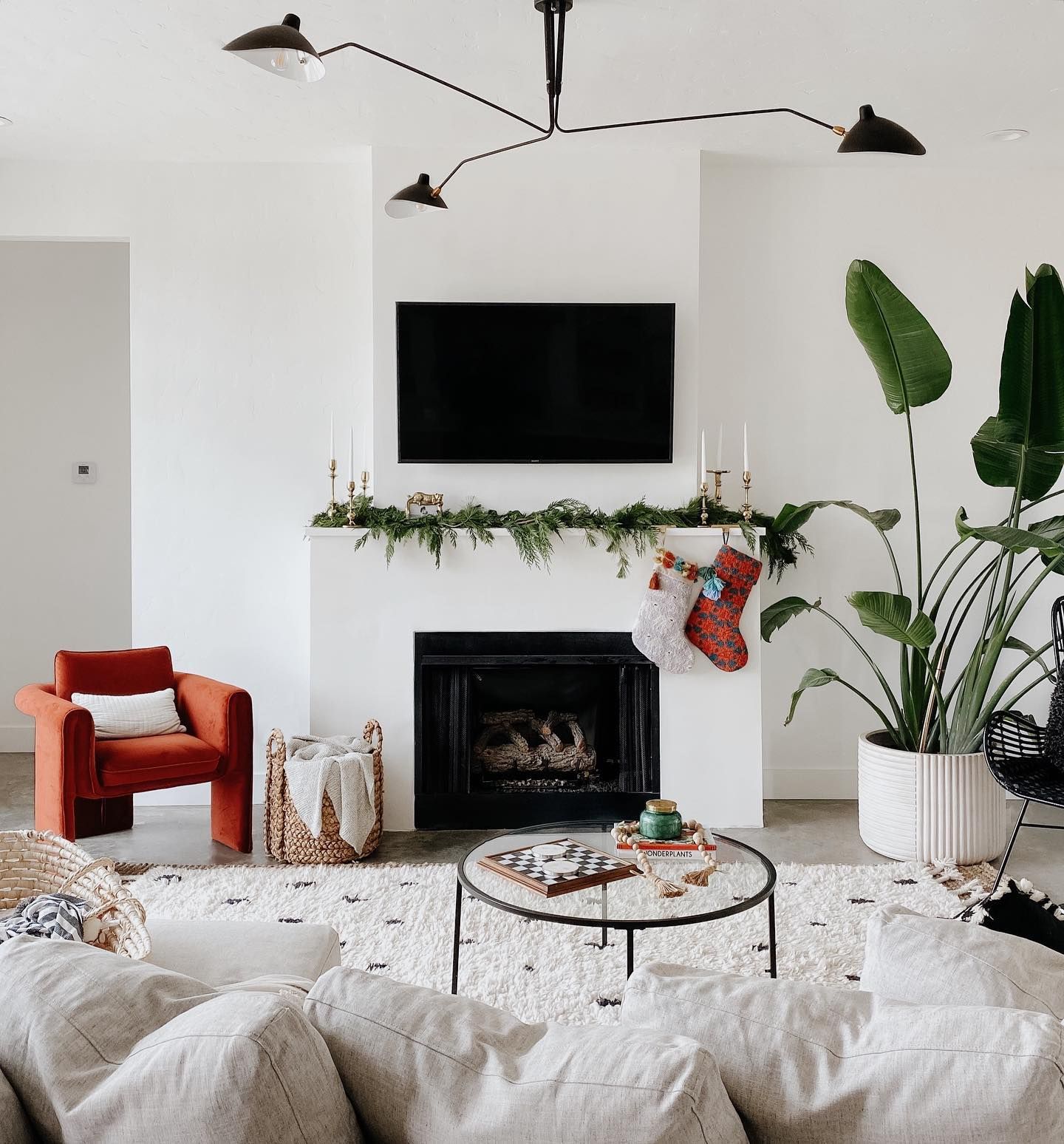 A modern living room with a fireplace, a white rug, a burnt-orange armchair, a glass coffee table, and a large houseplant.