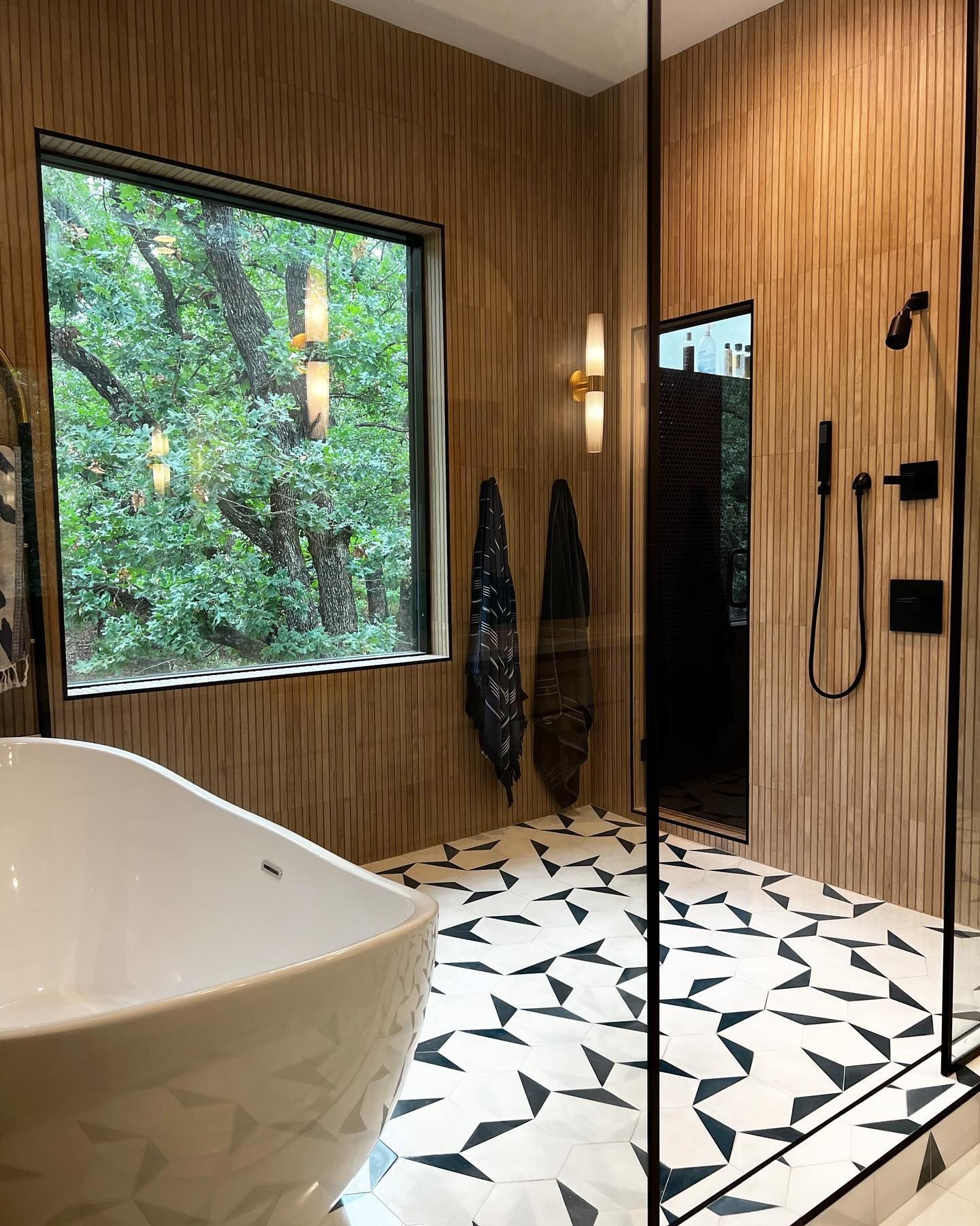 A modern bathroom with a freestanding white tub, geometric black and white tile, and wood-paneled walls looking onto trees.