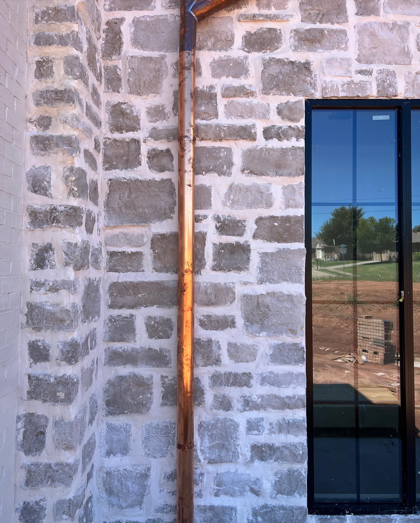 A copper downspout runs vertically against an exterior wall of light-colored, rustic stone next to a black-framed window.