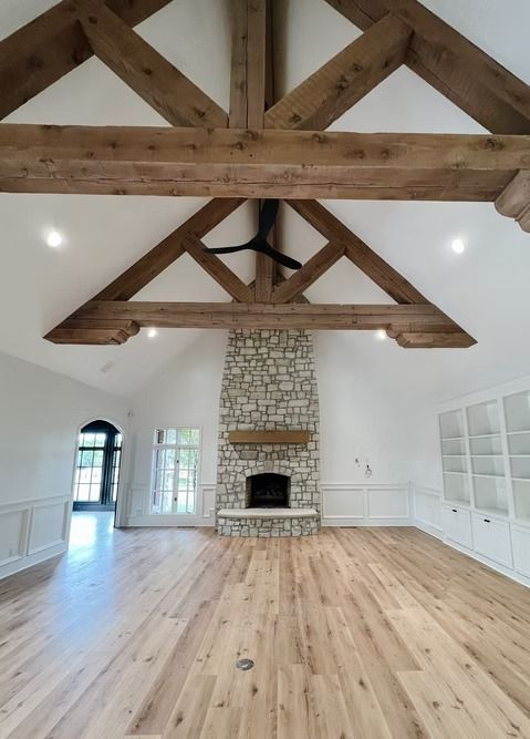 A spacious room with a vaulted ceiling, exposed wooden beams, light wood floors, and a floor-to-ceiling stone fireplace.
