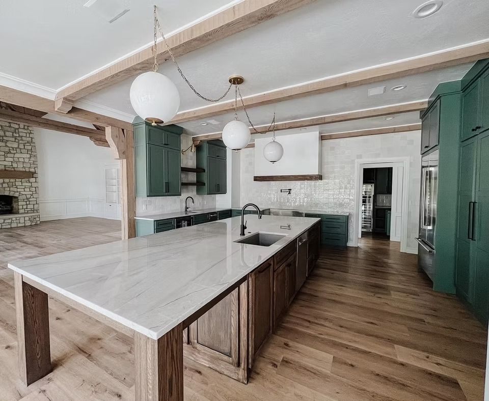 Modern kitchen with a long white stone island, wooden base, green cabinetry, and wood beam ceiling over light wood floors.