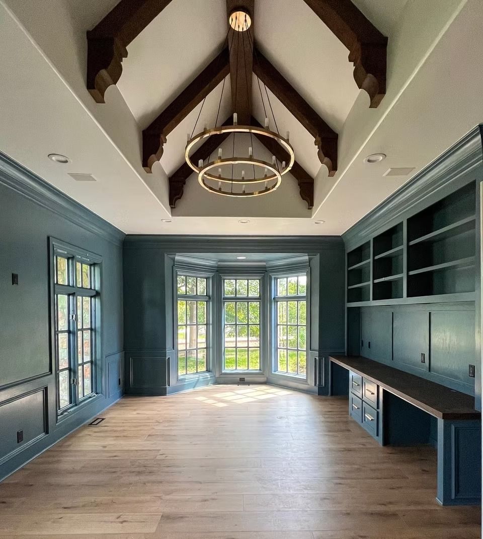 A home office with blue built-in shelves, wood floors, a vaulted ceiling with exposed beams, and a bay window.
