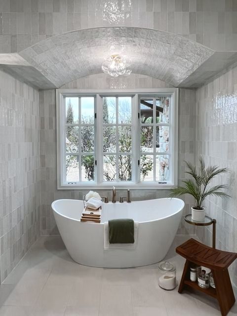 A white freestanding bathtub sits under an arched ceiling with a window view, beside a small plant and a wooden stool.