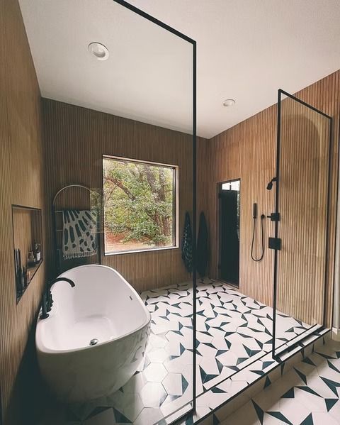 A modern bathroom features wood-paneled walls, a white freestanding tub, and a shower area with a geometric tile floor.