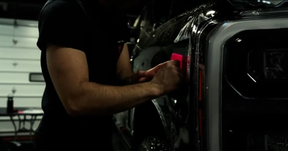 Person applying film to a vehicle's front side with a squeegee. Garage setting.