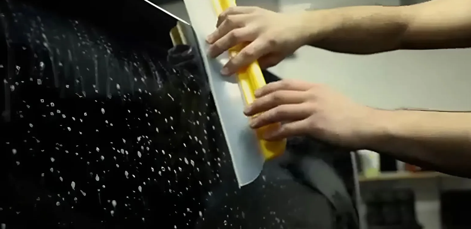 Hands applying a film to a black car window using a yellow squeegee.