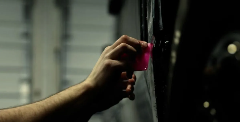 Person's hand using a pink squeegee on a black surface in a garage setting.