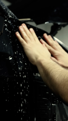 Hands applying soapy water to a black vehicle panel.