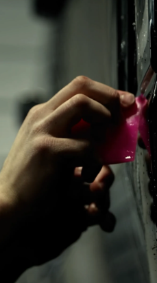 Hand using a pink squeegee on a dark, wet surface, possibly a vehicle window.