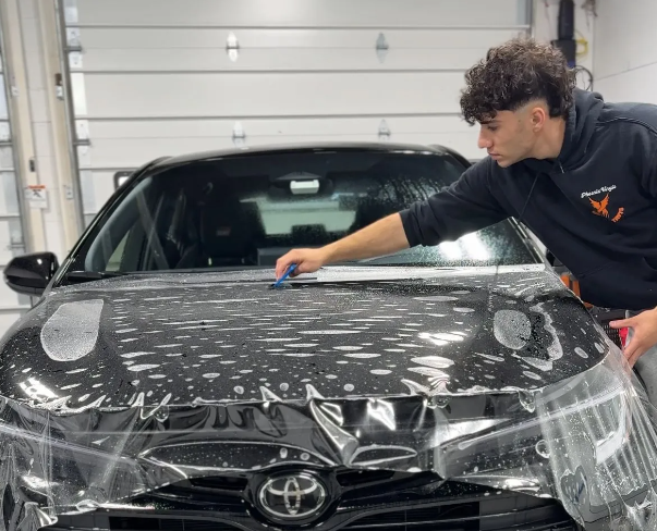 Man applying film to a black Toyota car hood. Interior shot, blue squeegee, professional setting.