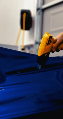 A person using a yellow infrared thermometer on a shiny blue car hood in a garage.