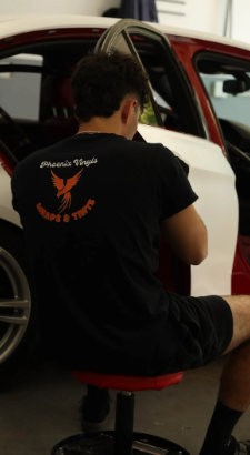 Person applying vinyl to a car door, wearing a black shirt with a phoenix logo, sitting on a red stool.