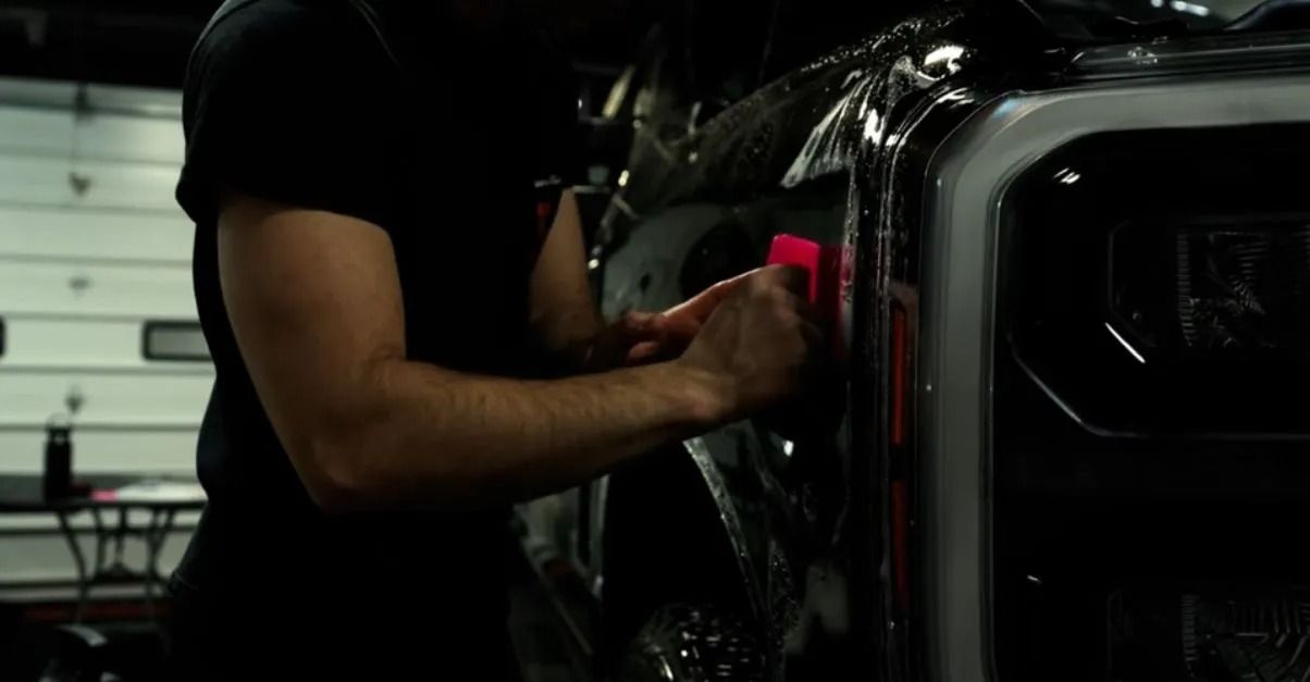 Person applying protective film to the front of a vehicle with a squeegee in a shop.