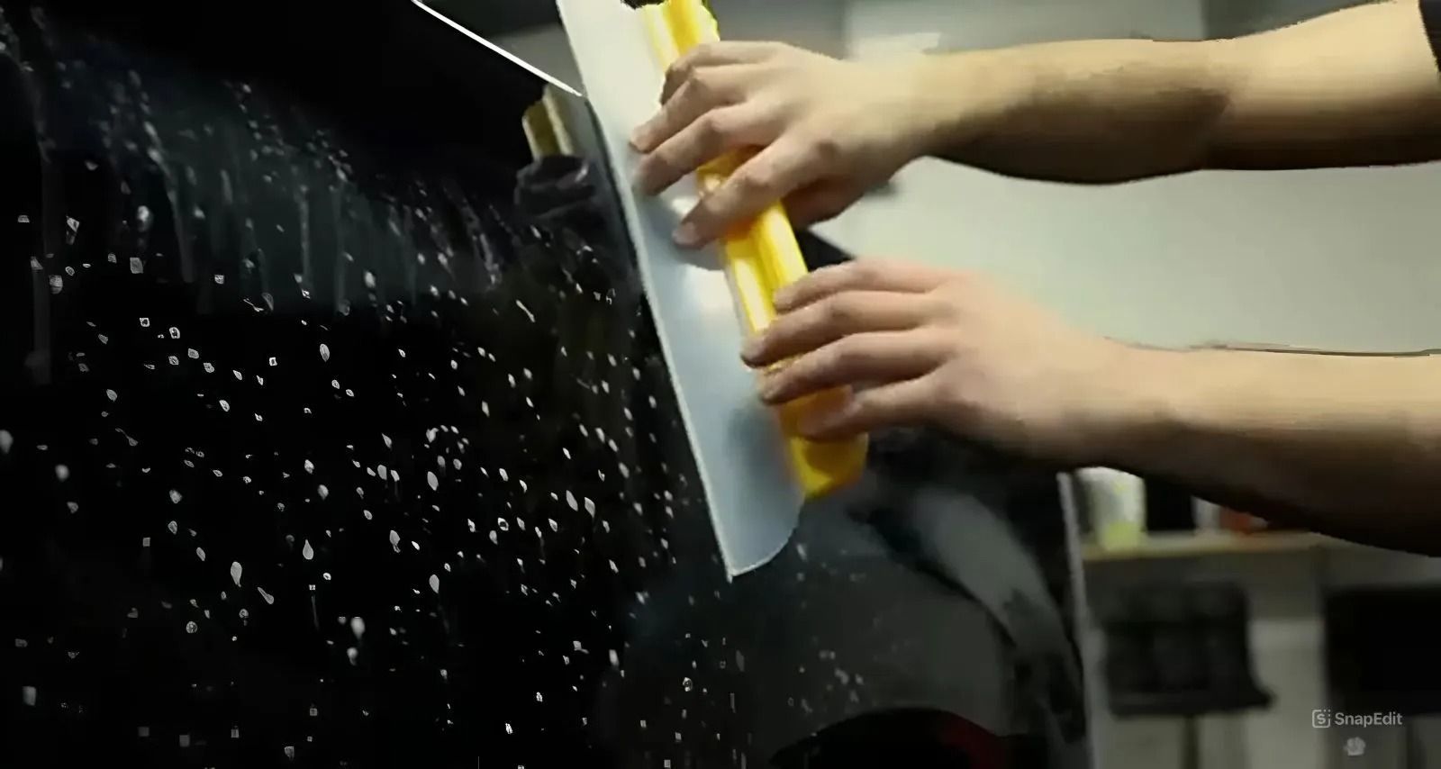 Person applying tint to a car window with a squeegee, soapy water visible, in an indoor setting.