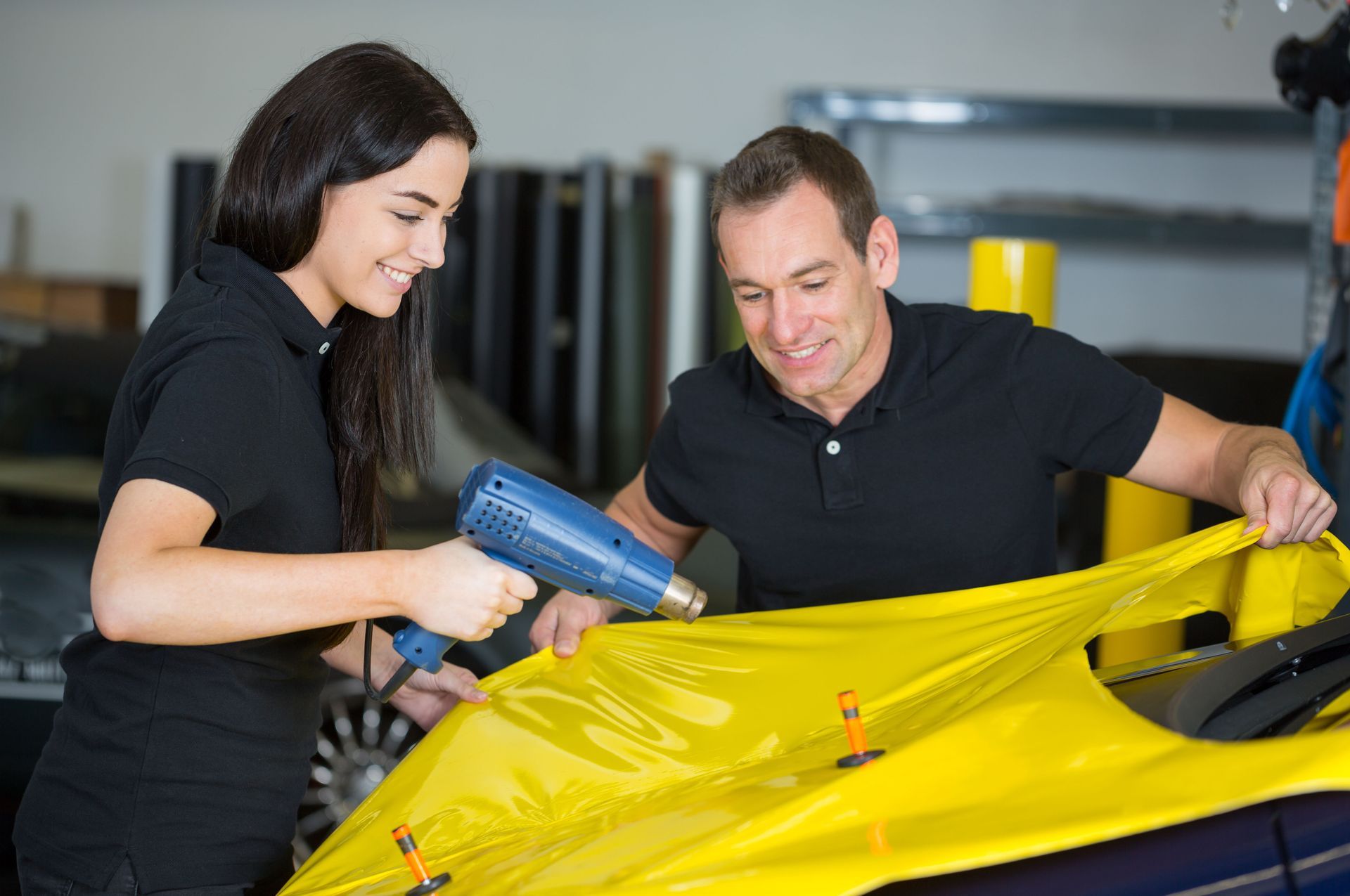 Two people using a heat gun to apply yellow wrap to a car hood in a garage.