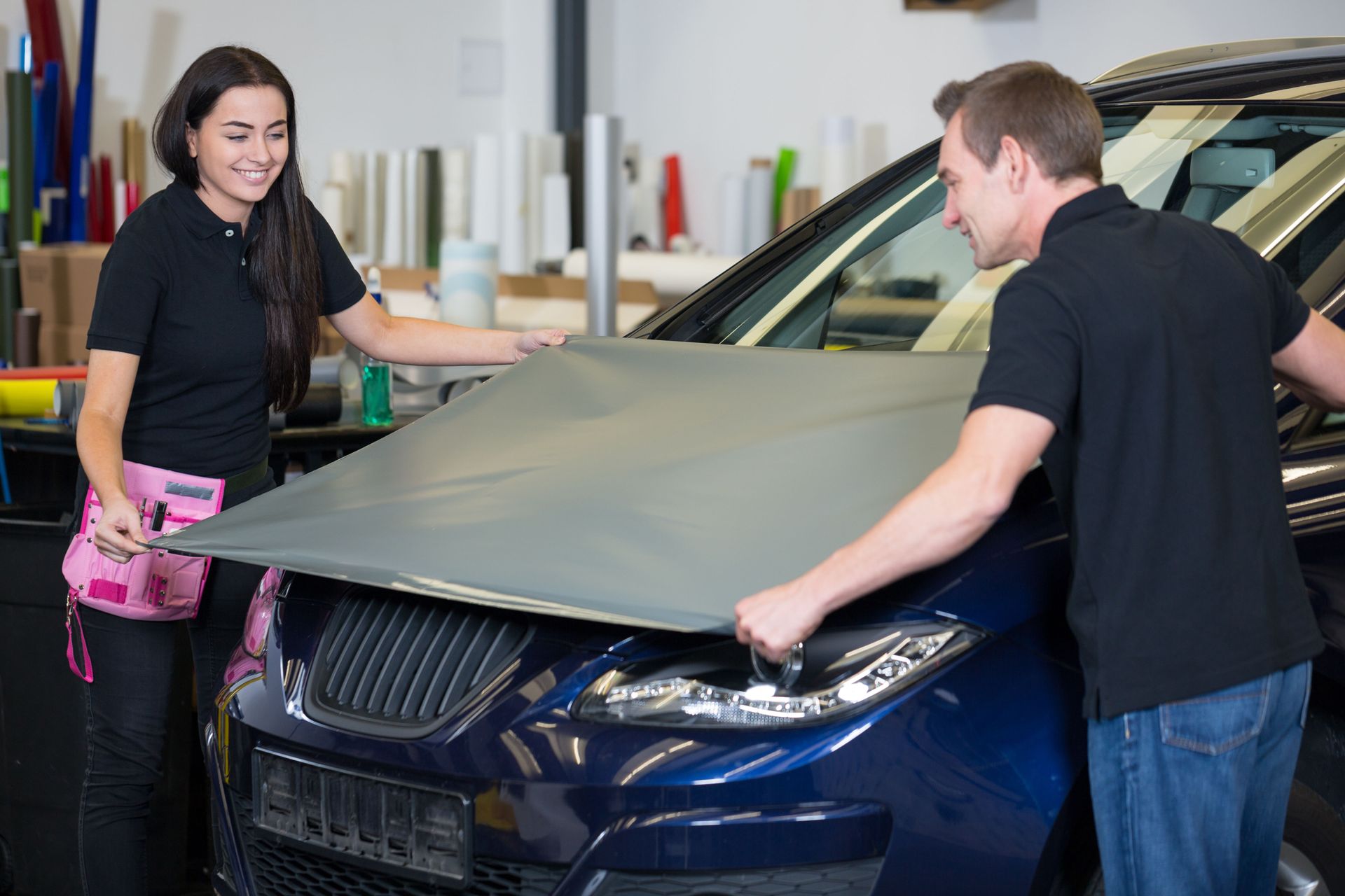 Two people applying gray vinyl wrap to the hood of a blue car in a shop.