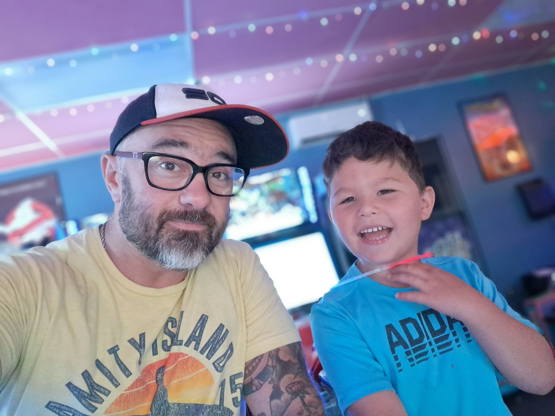 Man in baseball cap and child smiling, indoors, likely at an arcade.