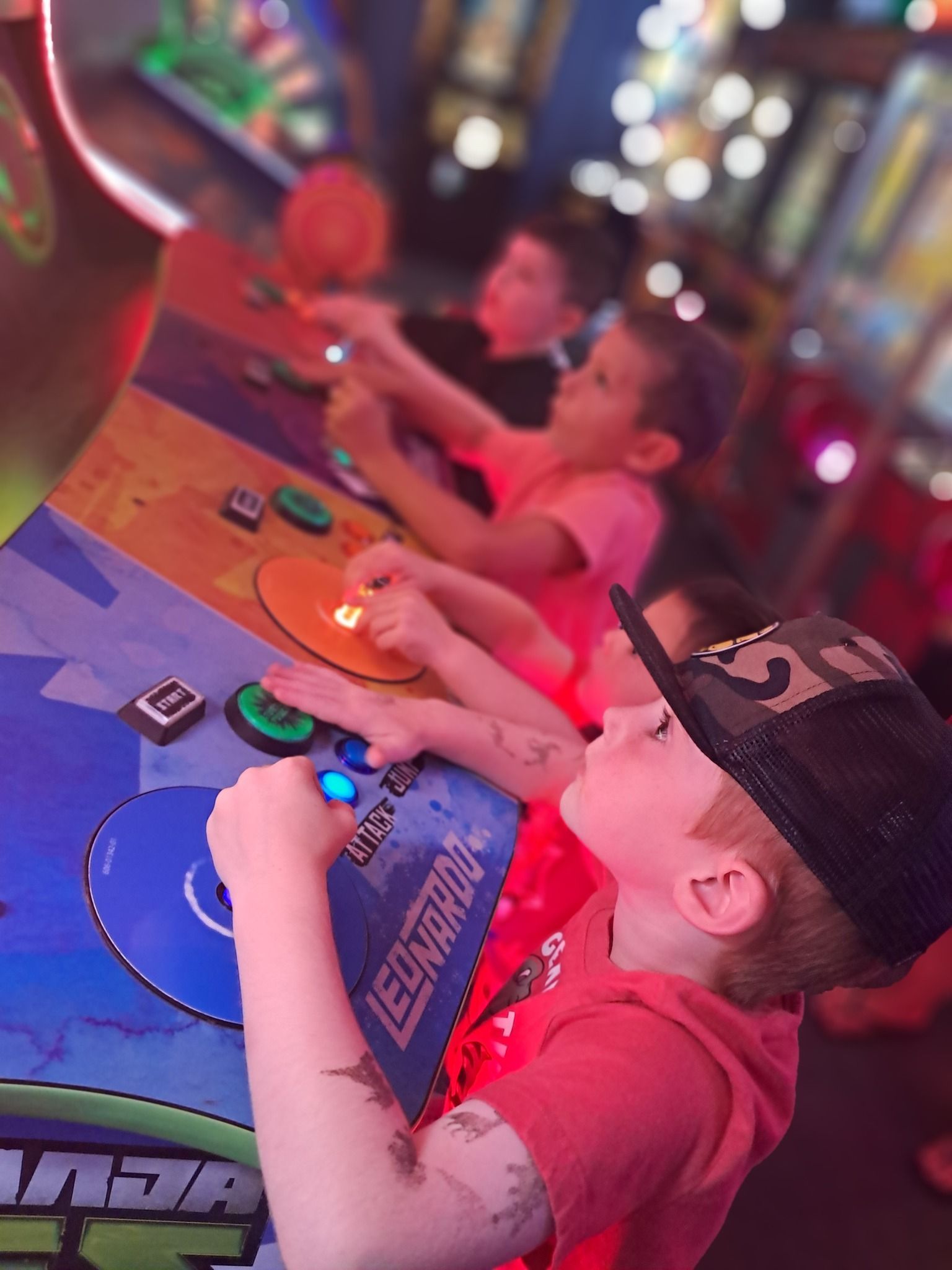 Children playing an arcade game. Three kids focused on colorful buttons. One in a hat, all wearing red shirts.