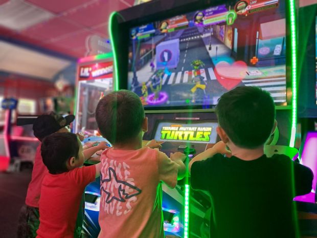 Kids playing Teenage Mutant Ninja Turtles arcade game; neon green lights, colorful screen, focused on game.