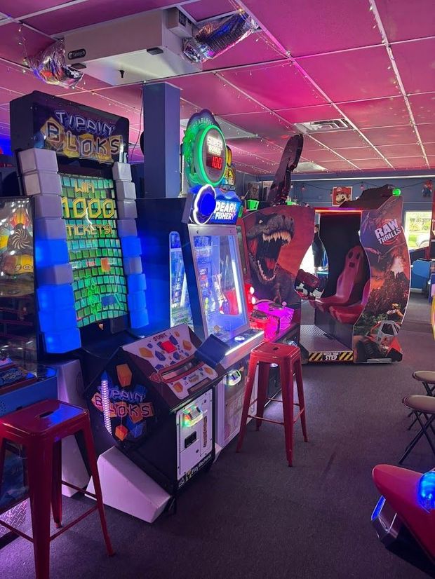 Arcade with glowing game machines, red stools, and pink lighting.