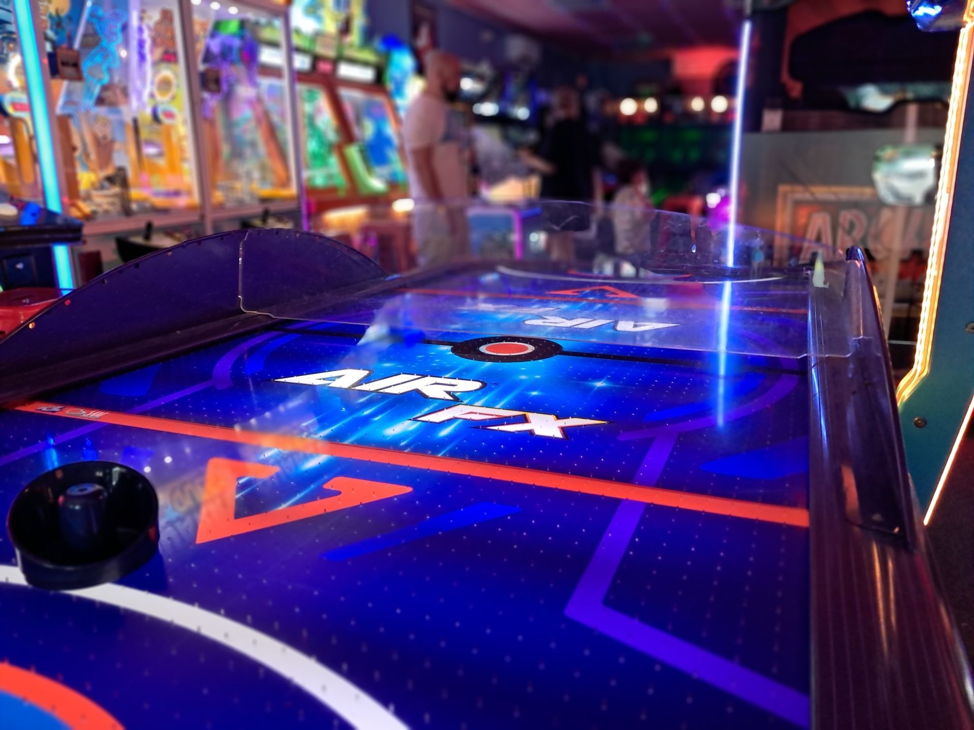 Air hockey table in an arcade with blurred figures in the background. Table is blue with neon accents.