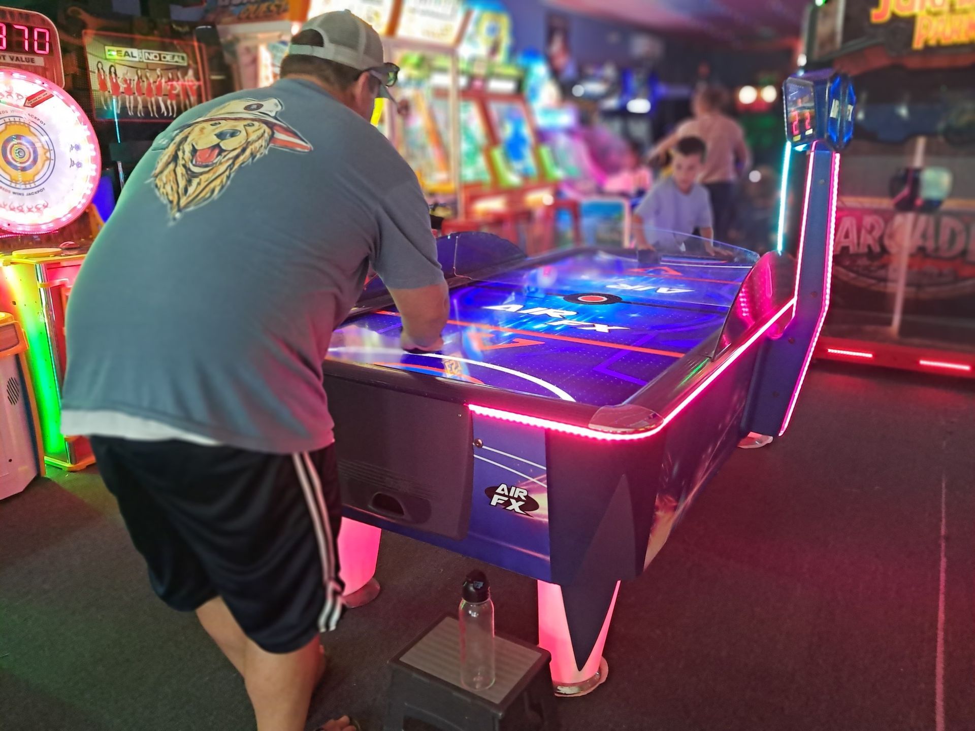 Man playing air hockey in an arcade, another person in background. Game table lit with neon lights.