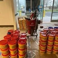 Stacks of red lidded containers sit on a plastic-covered floor, with a red rolling disposal bin and boxes in the background.