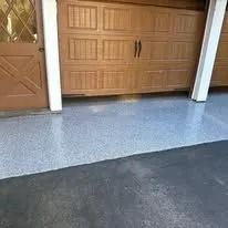 A light grey, speckled epoxy garage floor sits next to a dark asphalt driveway, leading to a wooden garage door.