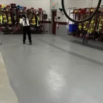 A person in uniform mops a smooth, gray floor in a fire station with rows of gear hanging on the wall.