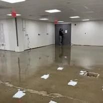A person stands in a flooded office space with reflective water covering the concrete floor and paper debris scattered.