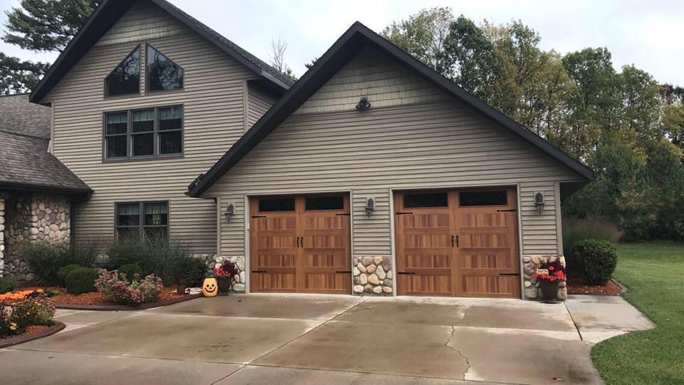 Two-story house with attached garage, tan siding, brown garage doors, and landscaped yard. Overcast sky.