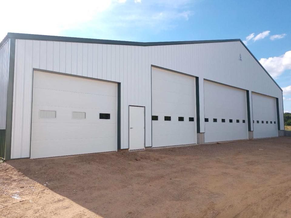 White industrial building with three garage doors and a small door, on a brown, dirt lot. Blue sky visible.