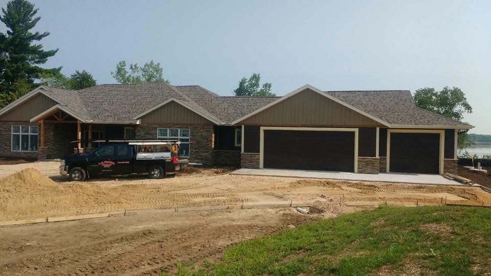 New house under construction, brown siding, two-car garage, truck parked in front, with lake in the background.