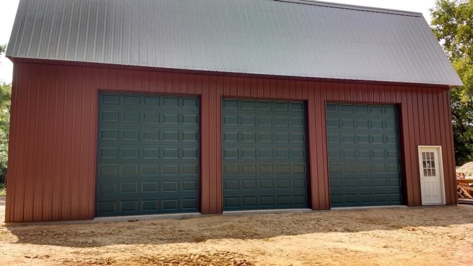 Red barn with three green garage doors and a white side door. Metal roof, gravel base.