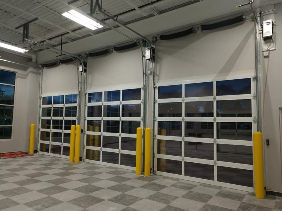 Three large, white garage doors with glass panels, yellow safety bollards, in a building with checkered flooring.