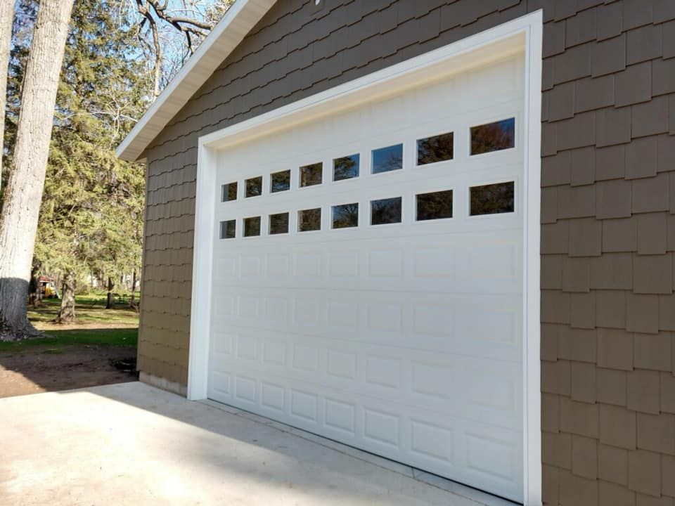 White garage door with square windows, set in a brown-sided building with a concrete driveway.